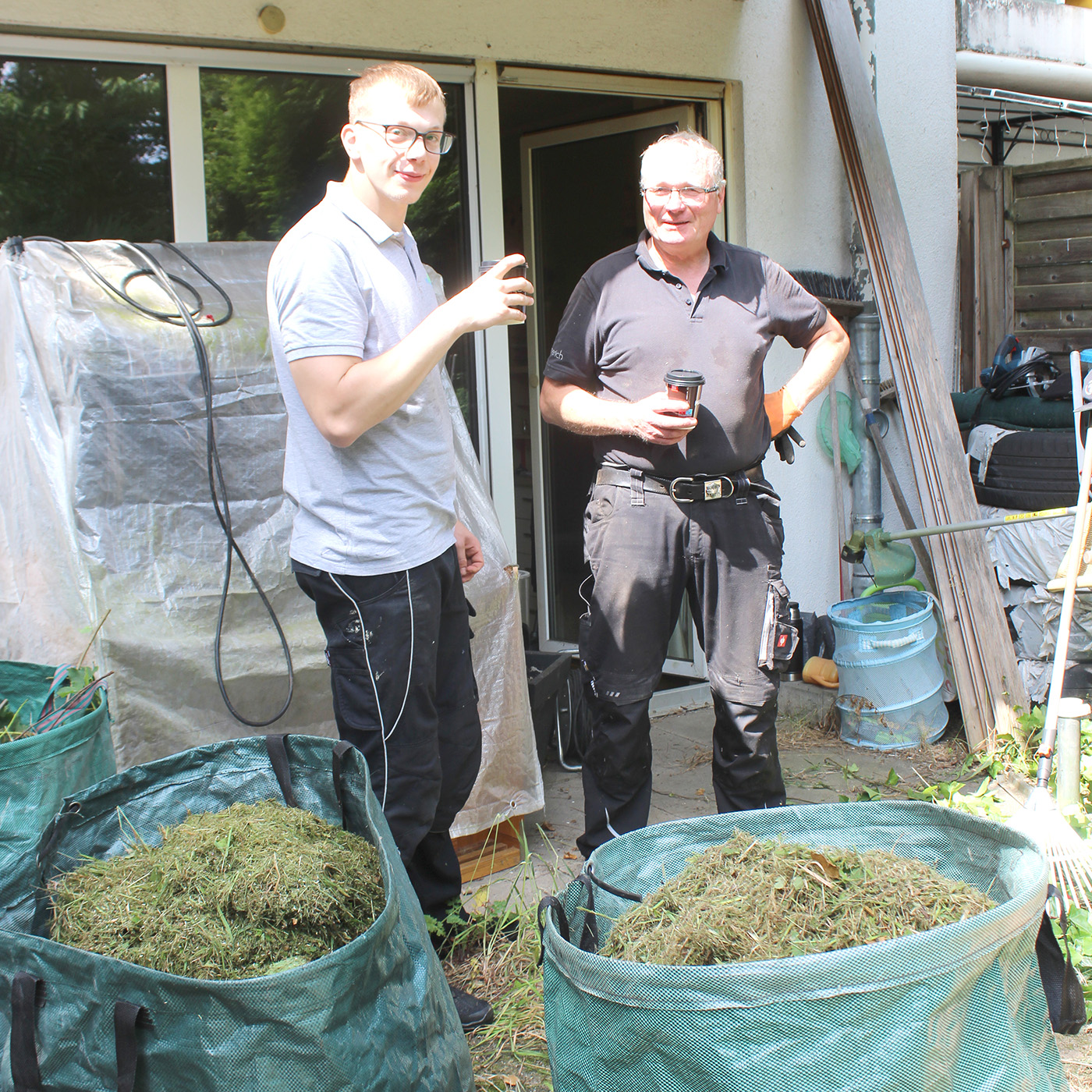 Ein älterer Herr und ein junger Mann stehen vor einem Haus. Vor Ihnen liegen Säcke mit Gartenabfällen. Sie erholen sich von der gemeinsamen Gartenarbeit.