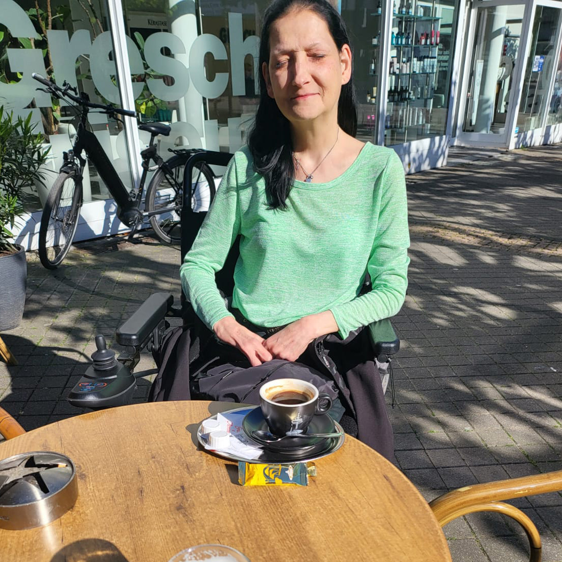 Eine Frau sitzt im Rollstuhl an einem Tisch eines Cafès. Sie genießt die Sonne und einen Kaffee.