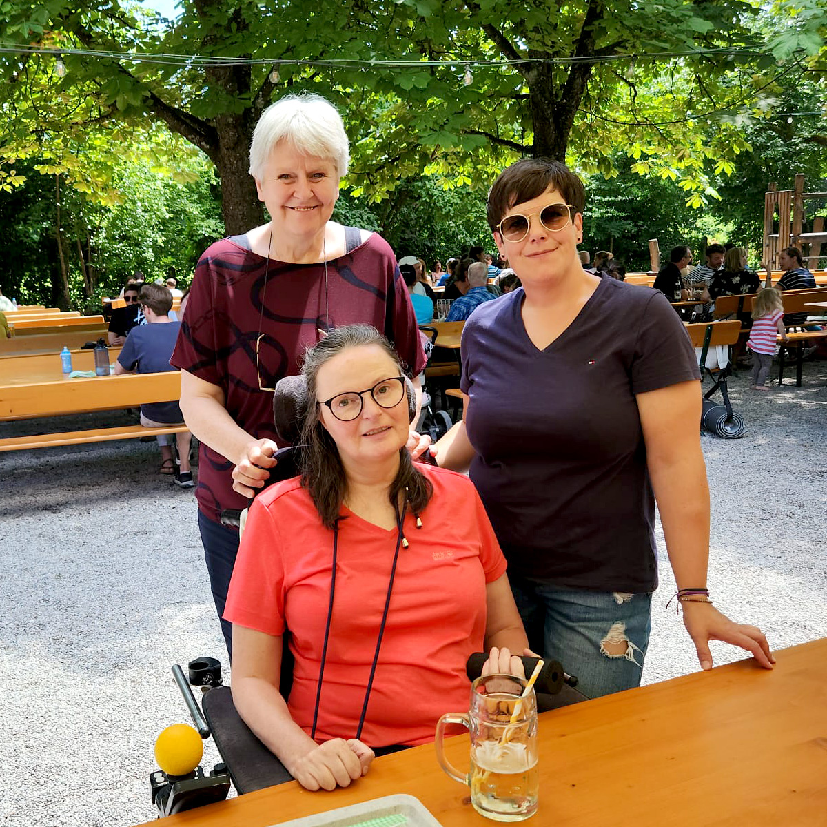 Drei Frauen befinden sich in einem Biergarten. Eine von Ihnen sitz in einem elektrischen Rollstuhl.