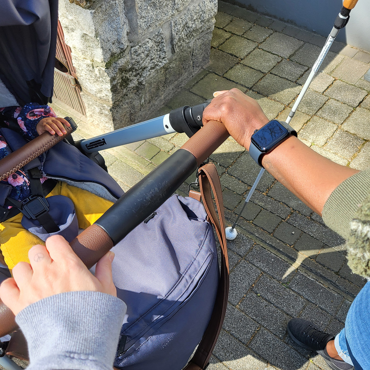 Zwei Frauen schieben gemeinsam einen Kinderwagen. Man sieht die Hände gemeinsam auf dem Griff ruhend. Eine der Frauen hält einen Blindenstock.
