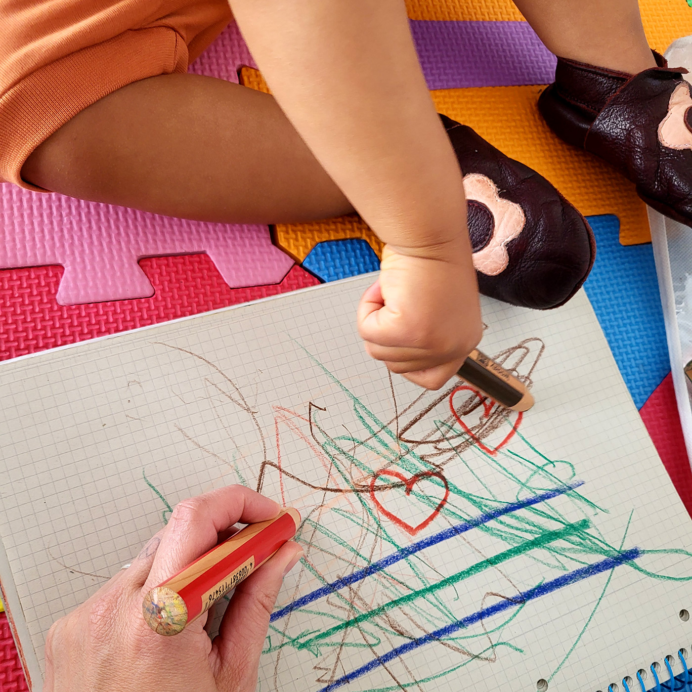 Ein kleines Mädchen malt mit Buntstiften - gemeinsam mit einer Kita-Assistenz - auf einem Blatt. Sie sitzen gemeinsam auf einem Kinderfussboden.