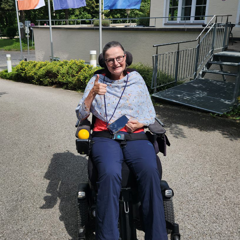 Eine Dame sitzt in Ihrem elektrischen Rollstuhl vor einem Ausflugscafé. Sie lächelt und macht eine 'Daumn hoch'-Geste. Sie schaut zufrieden.