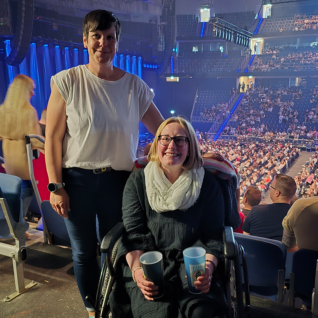 Zwei Frauen bei dem Besuch eines Musicals. Eine der Frauen sitzt im Rollstuhl und lächelt. Sie hält zwei Getränkebecher. Neben Ihr steht Ihre Assistentin - passend für den Anlass gekleidet.