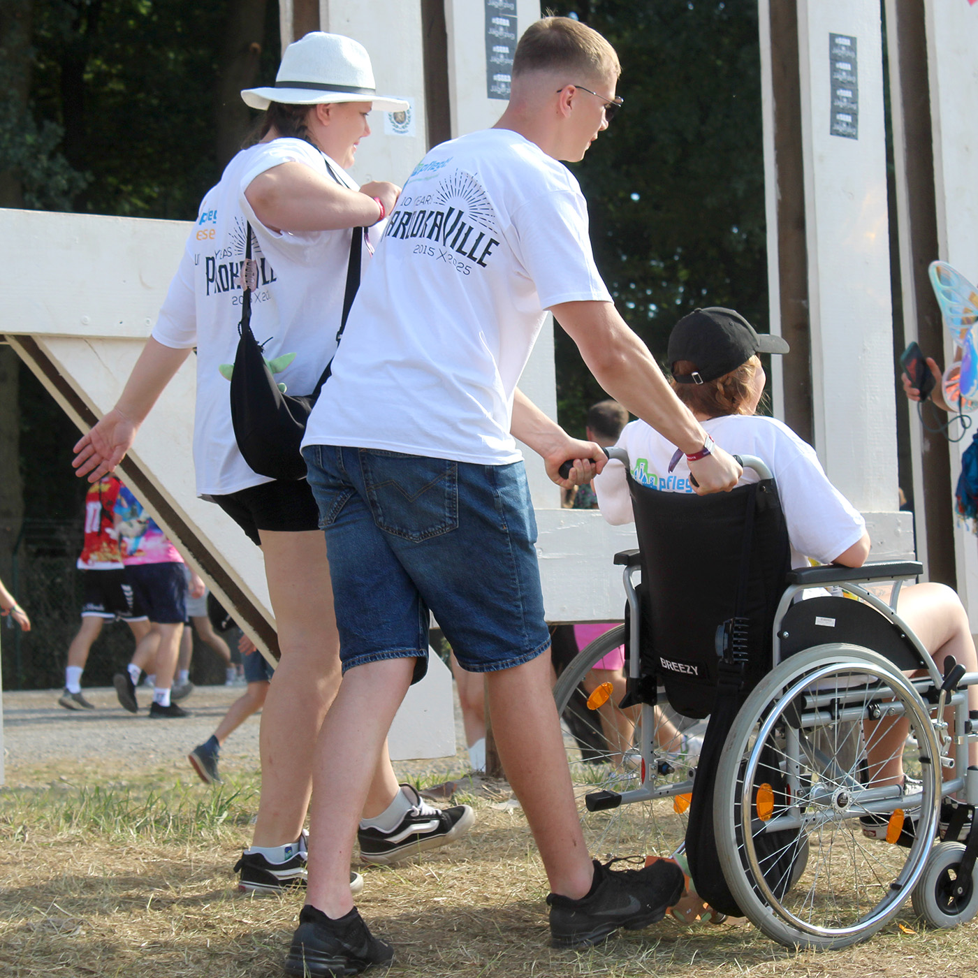 Eine junge Frau im Rollstuhl wird auf einem Festivalgelände von zwei Assistenten begleitet.
