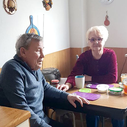 Ein langjähriges Ehepaar sitzt gemeinsam am gedeckten Frühstückstisch und unterhält sich.