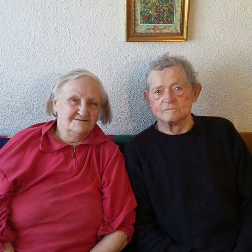 Ein altes Ehepaar sitzt auf dem Sofa in Ihrer Wohnung. Beide lächeln in die Kamera.