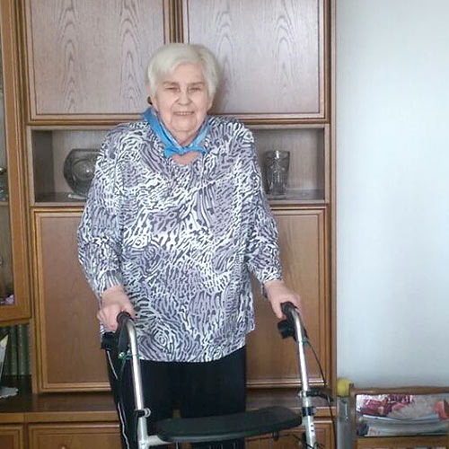 Eine alte Dame mit Rollator steht in Ihrem Wohnzimmer und lächelt.