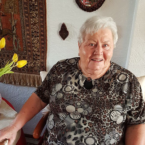 Eine rüstige Dame sitzt an Ihrem Wohnzimmertisch und schaut freundlich in die Kamera.
