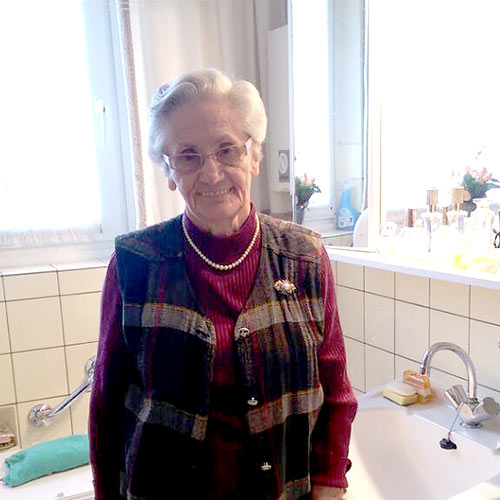 Eine alte Dame steht am Waschbecken Ihres Badezimmers. Sie ist adrett gekleidet und lächelt in die Kamera.