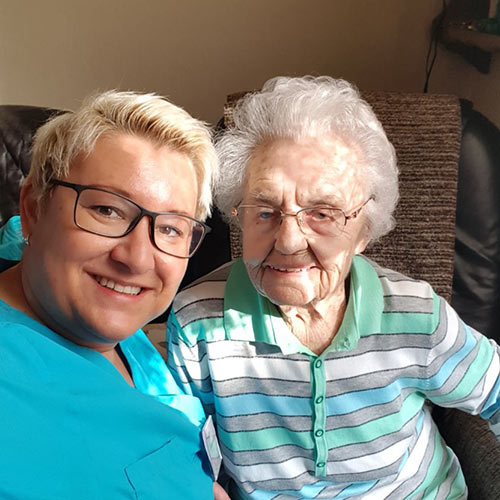 Eine alte Dame sitzt gemeinsam mit einer Pflegedienst-Mitabeiterin auf der Couch. Beide Lächeln in die Kamera.