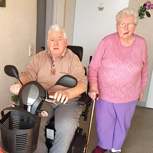 Ein Mann mit einer elektrischen Gehhilfe sitzt im Eingangsbereich seiner Wohnung. Neben Ihm steht seine Lebensgefährtin. Sie nutzt einen Gehstock.