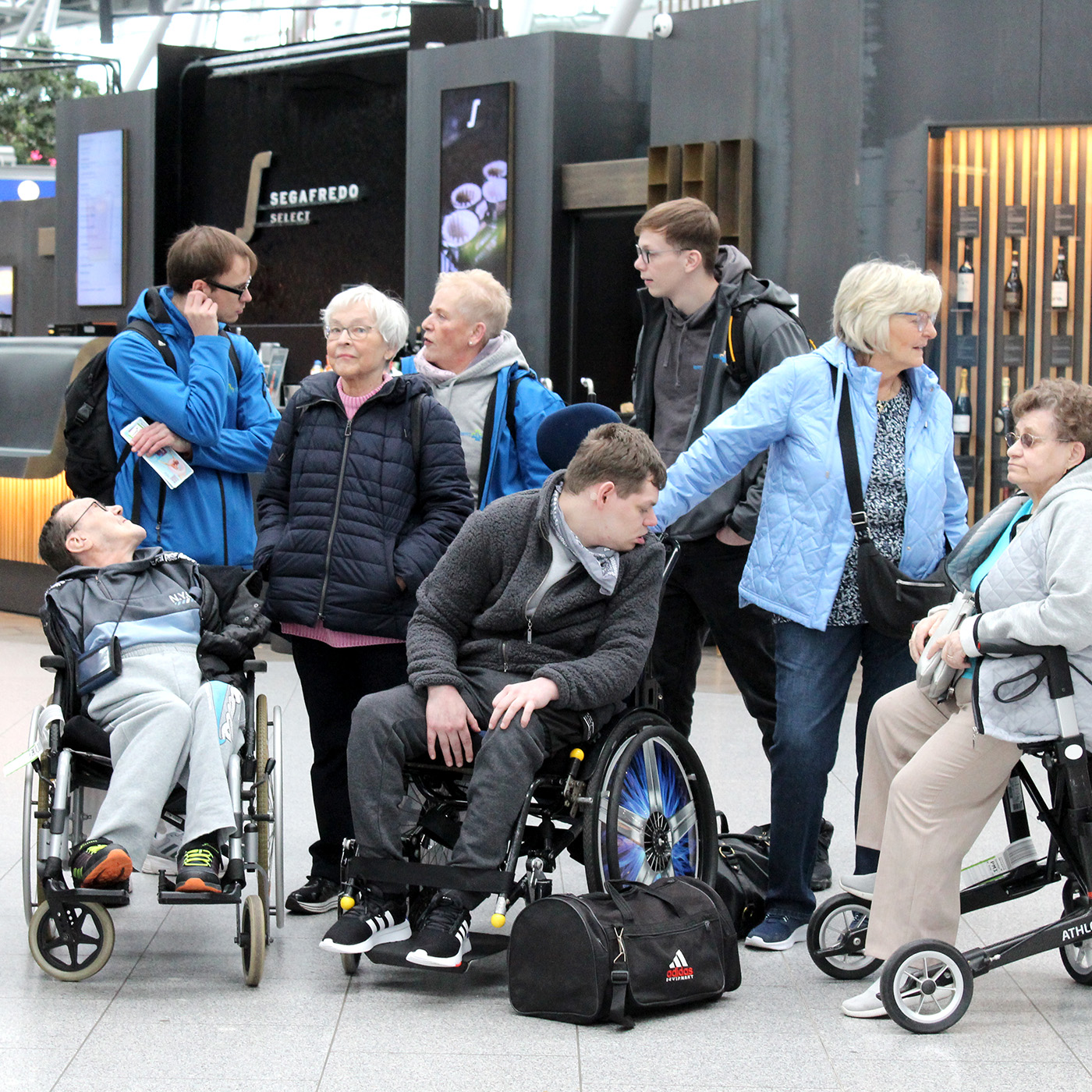 Eine WAT-pflegt! Reisegruppe wartet am Flughafen auf den Check-In. Die Reisegruppe besteht aus mehreren Menschen, mit unterschiedlichen Handicaps. Sie werden Begleitet von unserem Team.