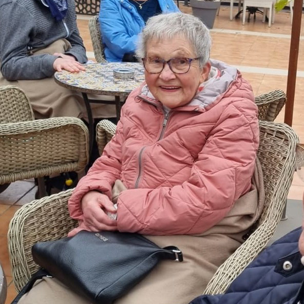 Eine alte Dame sitzt in einem Korbstuhl in einem Straßencafé. Sie lächelt.